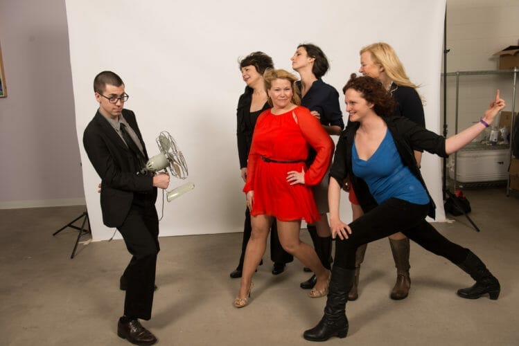 A group of six women in stylish outfits poses playfully around a man holding a vintage fan, set against a plain backdrop.