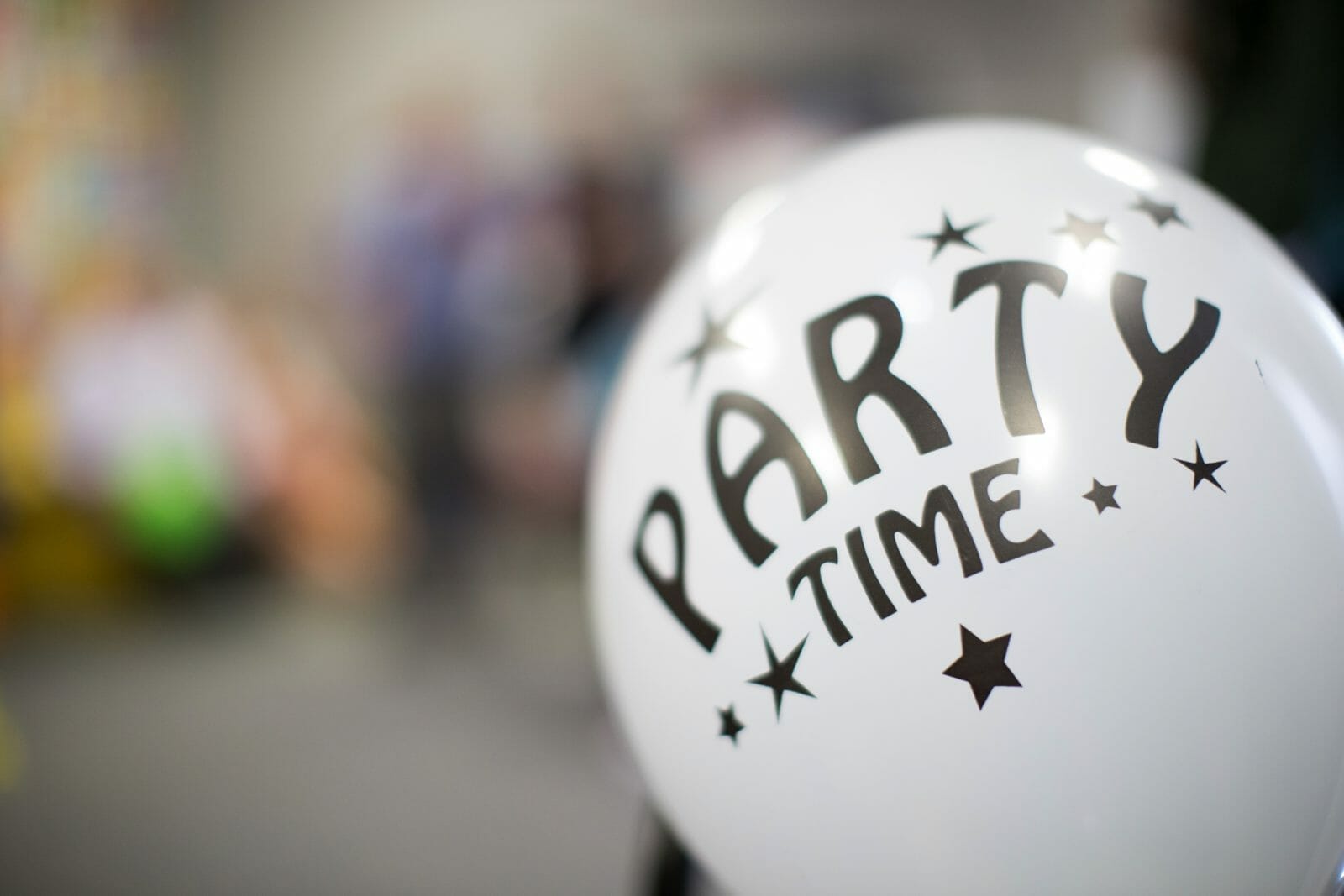 White balloon with "PARTY TIME" text and stars, set against a blurred festive background.