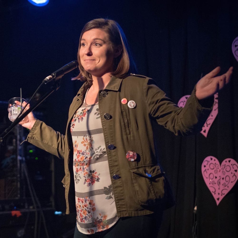 A woman in a floral top and green jacket gestures while holding a drink, speaking at a microphone with heart decorations in the background.