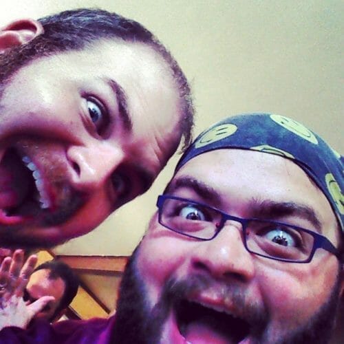 Two men making exaggerated facial expressions, smiling widely with playful excitement, wearing casual attire and accessories.