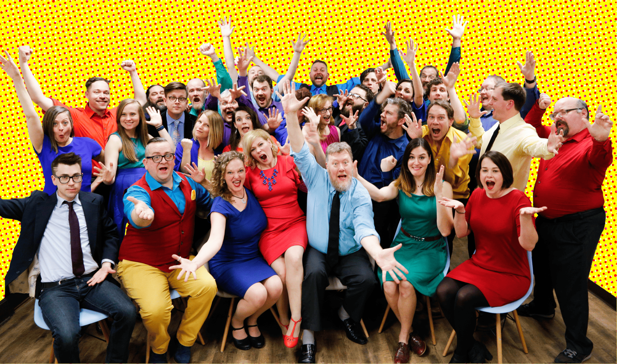 A large, diverse group of people in colorful outfits joyfully posing with raised hands against a bright yellow dotted background.