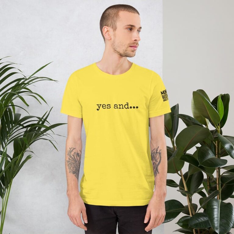 A man with tattoos wears a bright yellow t-shirt featuring the phrase "yes and..." against a backdrop of greenery.