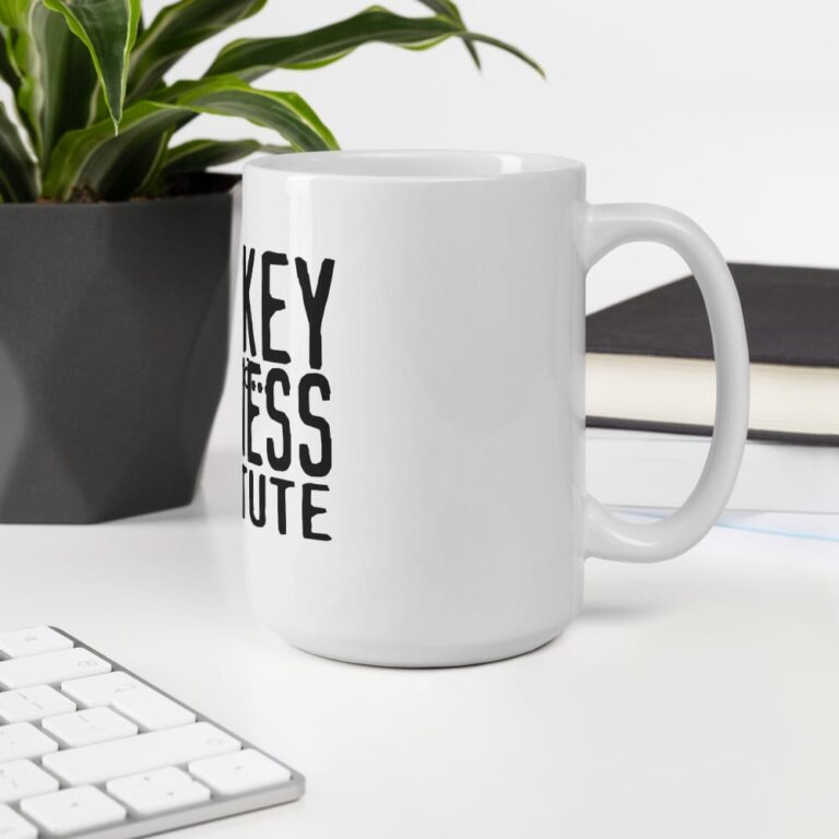 White mug with bold black text beside a potted plant and a keyboard on a clean desk surface.