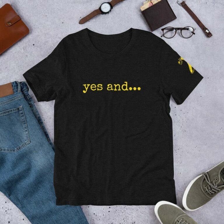 Black t-shirt with "yes and..." in yellow, paired with blue jeans, gray sneakers, and accessories on a gray background.