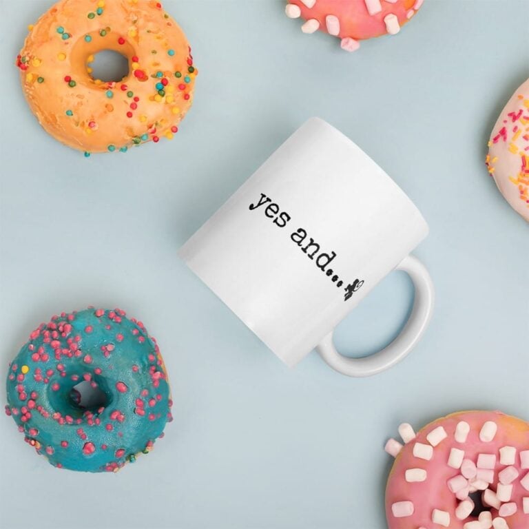 A white mug with "yes and..." text surrounded by colorful donuts on a light blue background.