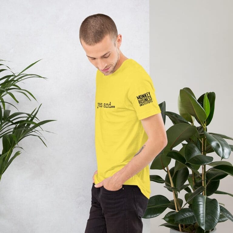 Young man in a yellow t-shirt with "yes and..." text, standing beside green plants against a light background.
