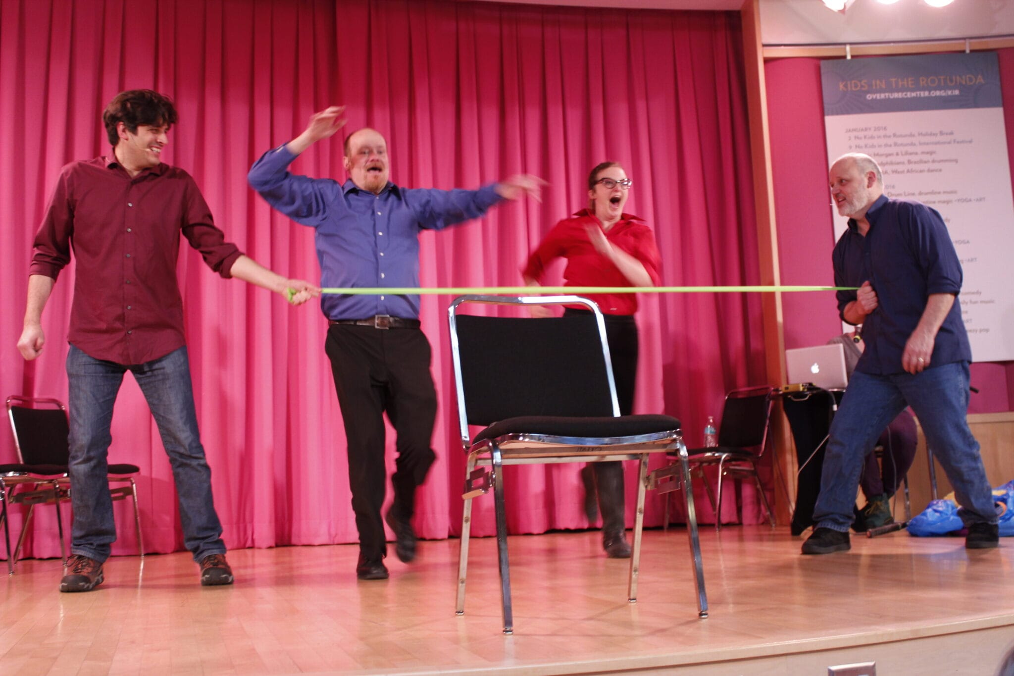 Four performers engage in a lively skit, pulling a yellow ribbon while a chair sits empty on stage with pink curtains behind.