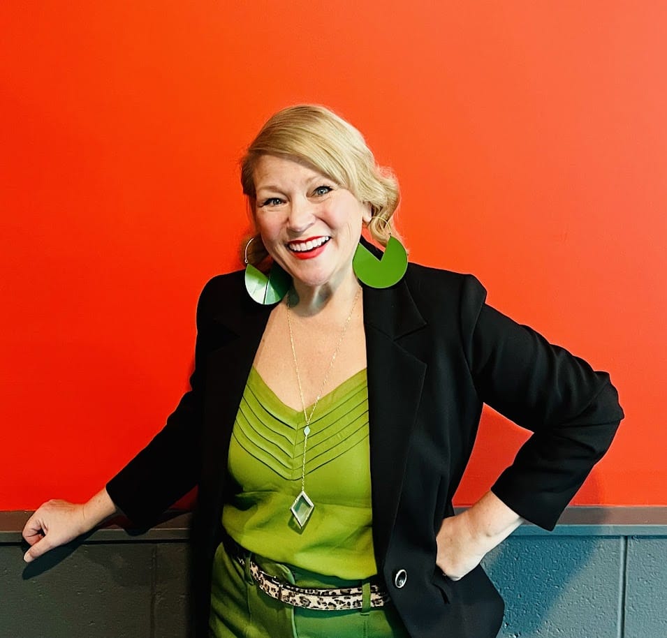Woman in black blazer and green top with large green hoop earrings smiles against an orange wall.