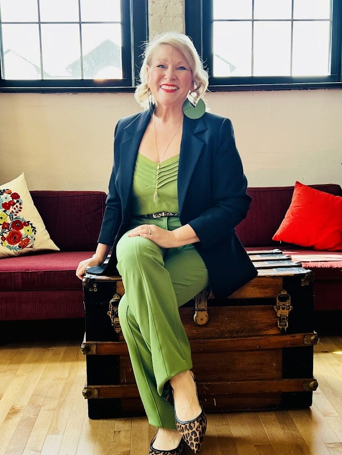 Woman in green suit and leopard heels sits on antique trunk.